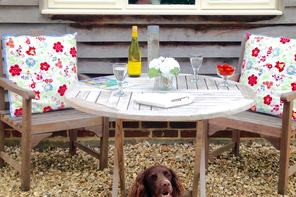 An outdoor seating area features a round wooden table surrounded by two benches adorned with floral cushions. A bottle of wine and glasses are displayed alongside a delicate flower arrangement. A friendly dog sits on the gravel beneath the table, while a window brings natural light to the space.