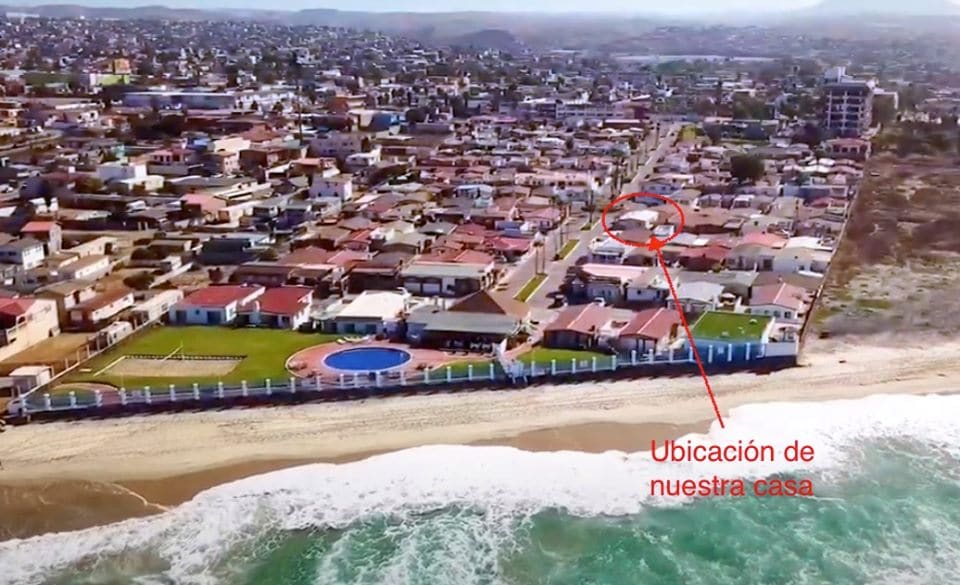 An aerial view captures the neighborhood along the coastline, showcasing a mix of residential homes and the beach. A shimmering pool is visible in the foreground, with the house location indicated. The landscape blends urban living with seaside charm.
