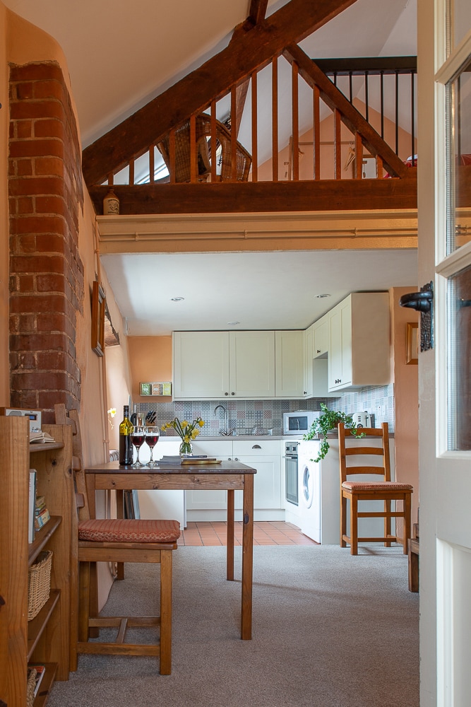 A cozy open-plan layout features a dining table with two chairs, accompanied by a well-equipped kitchen area showcasing white cabinetry. Warm tones on the walls complement the wooden beams that highlight the ceiling above, creating a welcoming atmosphere throughout the space.