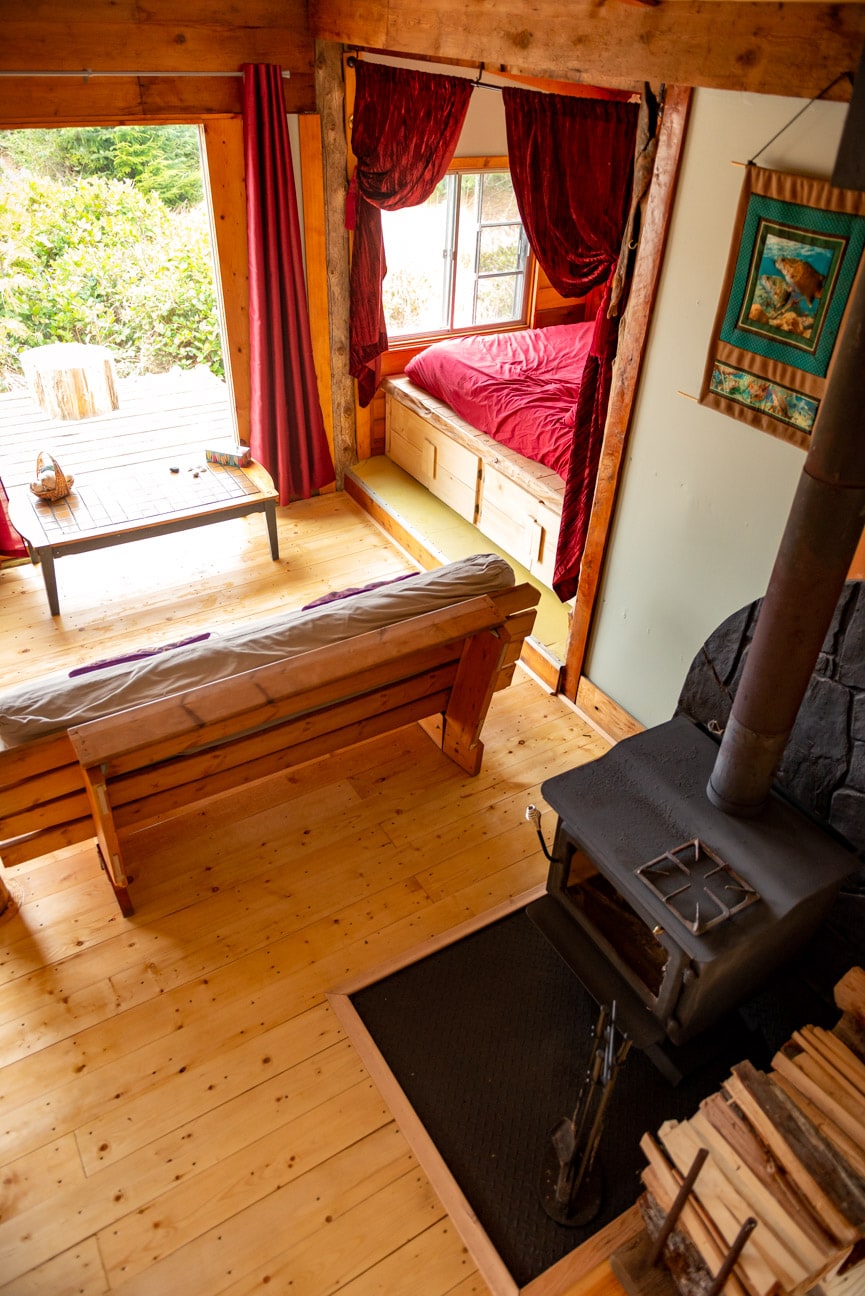 An overhead view of the cabin reveals a warm interior with wooden flooring. A cozy sofa is positioned near a small table. A red-curtained bay bedroom is visible in the background, complemented by a wood stove and neighboring wood storage.