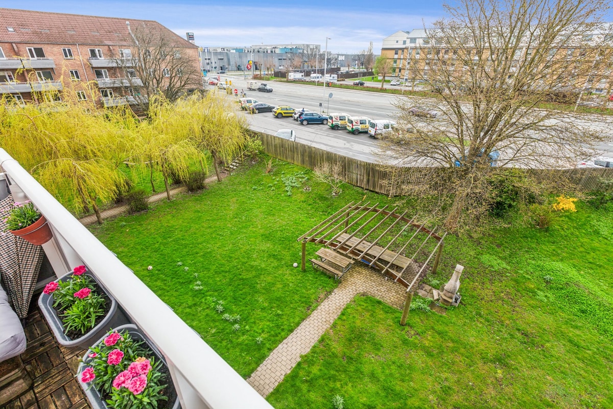An overview of the surrounding green space is provided, featuring a well-maintained lawn with a pergola and seating area. Flower pots with vibrant pink flowers are positioned on the balcony railing, enhancing the view towards the landscaped garden.