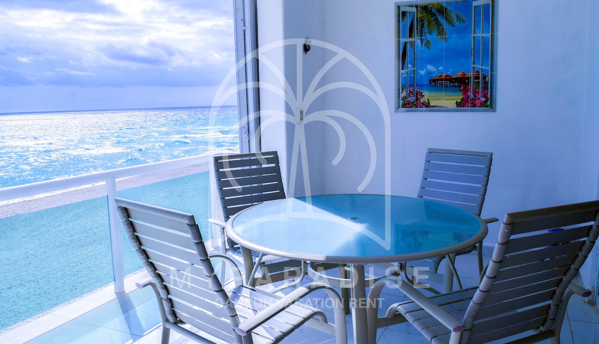 A round glass dining table is set on a large balcony, surrounded by six light-colored outdoor chairs. The ocean view stretches in the background, reflecting soft blue tones under a cloudy sky. A framed beach-themed artwork is visible on the wall nearby.