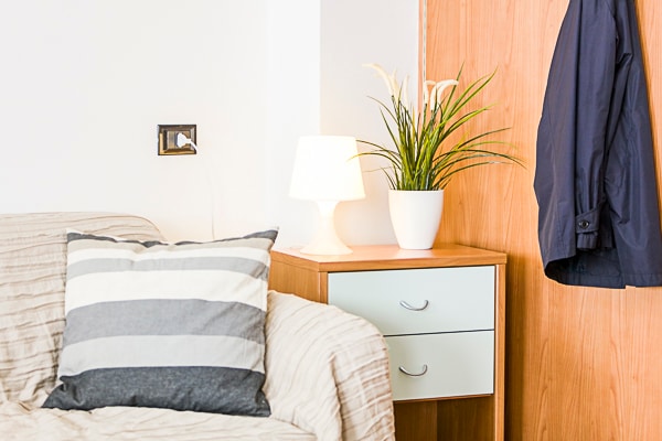 A neutral-toned living area is highlighted by a cozy sofa with striped cushions. A bedside table with two drawers features a potted plant and a soft lamp, providing additional warmth to the space. A jacket hangs casually on a wooden door nearby.