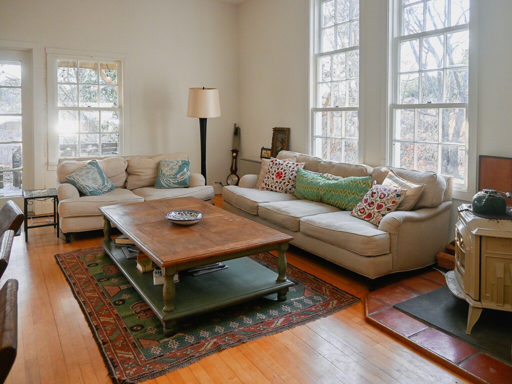 A spacious living area features large windows allowing natural light to fill the room. A comfortable sectional sofa is arranged around a wooden coffee table, which rests on a decorative area rug. A lamp and framed artwork complement the inviting space.