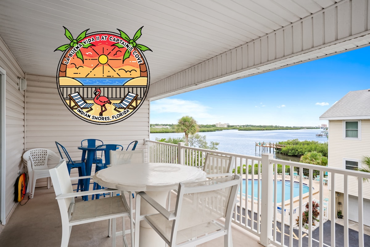 A private balcony features a round table with four white chairs, overlooking a serene view of the Intracoastal Waterway and swimming pool below. The clear sky enhances the tranquil atmosphere, complemented by the surrounding lush greenery.