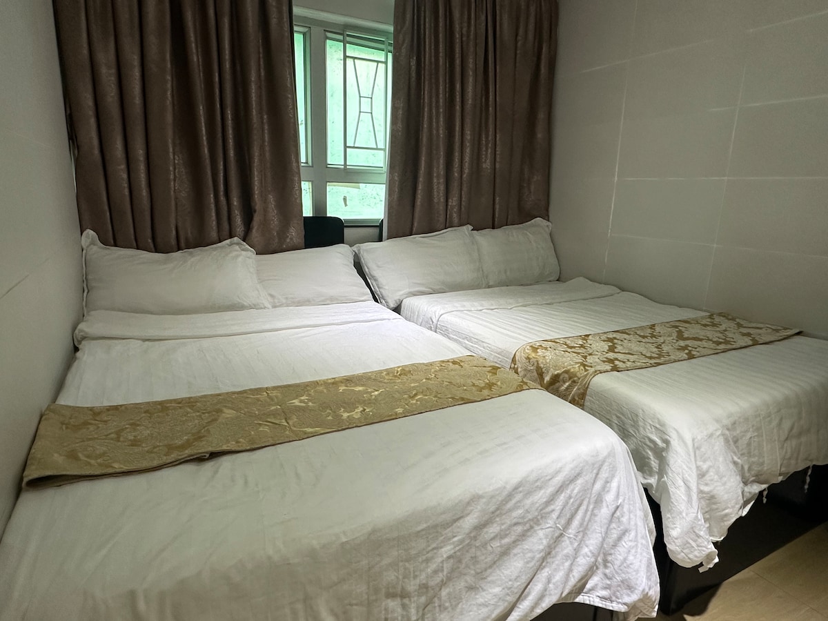 The room contains two spacious beds, each dressed in white linens with decorative gold accents. Soft curtains frame a window that provides natural light, creating a comfortable and inviting atmosphere.
