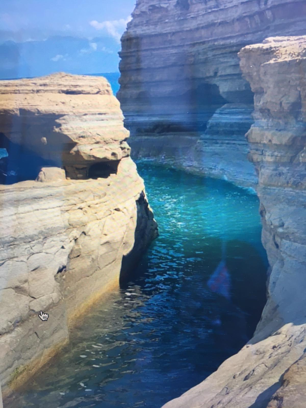 A rocky shoreline is depicted, featuring layered sandstone cliffs surrounding a serene turquoise water channel. The sunlight reflects off the water's surface, enhancing the tranquil atmosphere. The cliffs have unique formations, creating a picturesque natural environment.