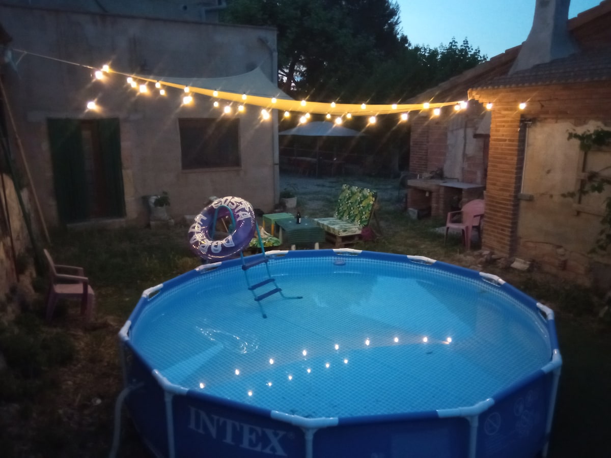 A circular inflatable pool is set in a grassy area, illuminated by string lights overhead. A colorful inflatable float rests in the pool, while a ladder leads into the water. Nearby, simple chairs are positioned around the area, contributing to a relaxed outdoor setting.