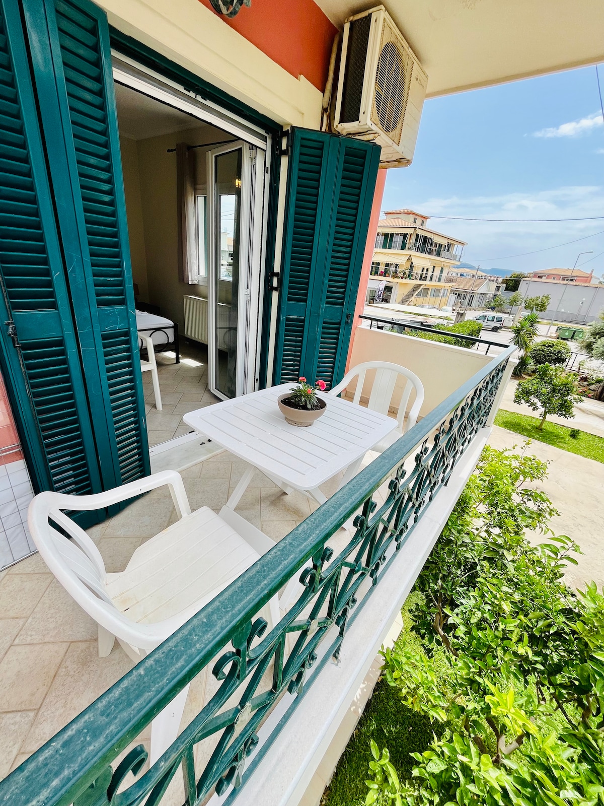 A balcony is presented with a small white table and two chairs, accompanied by a potted plant. The view includes a quiet street and a glimpse of neighboring buildings, enhancing the feeling of outdoor relaxation.