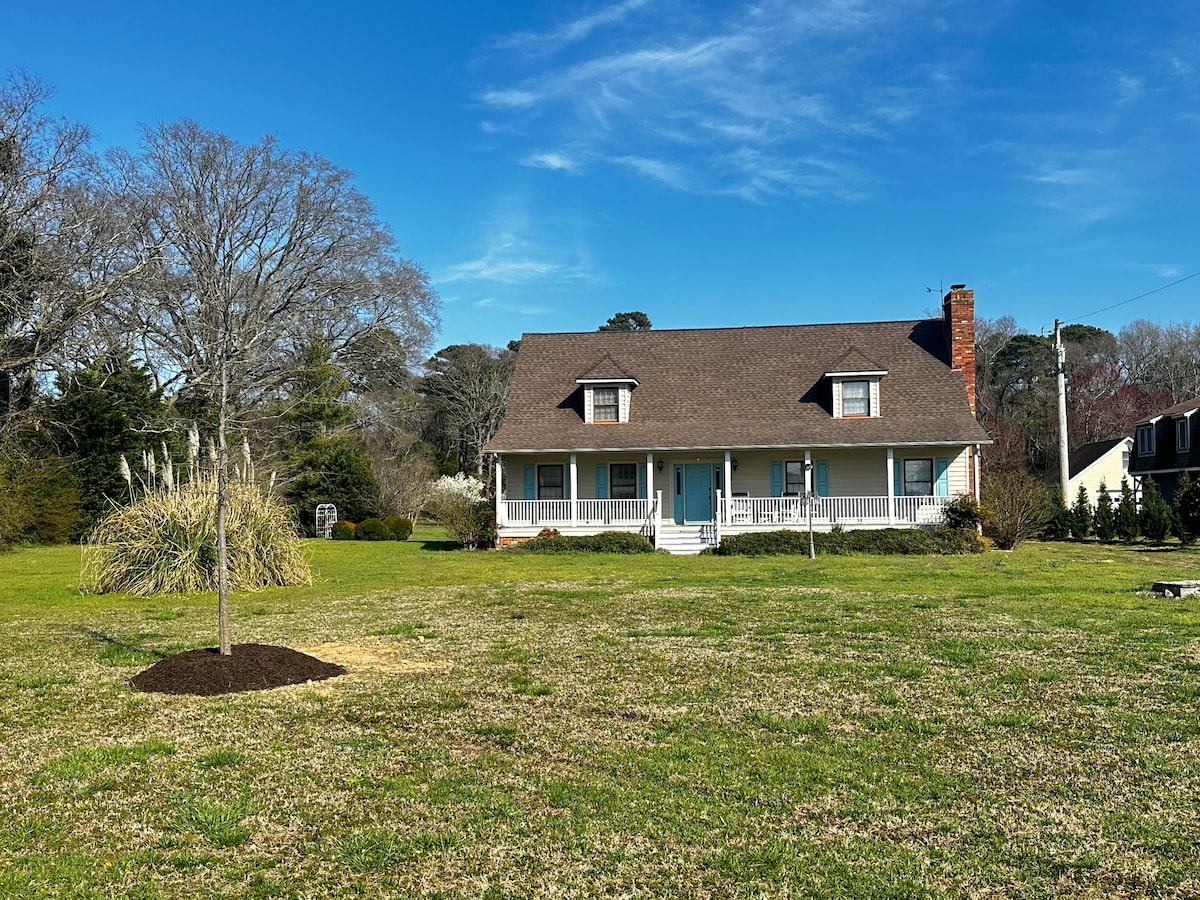 A two-story bungalow is shown against a clear blue sky, featuring a sloped roof and a welcoming front porch. A spacious lawn surrounds the home, with grassy areas and a young tree planted nearby, creating an open and inviting landscape.