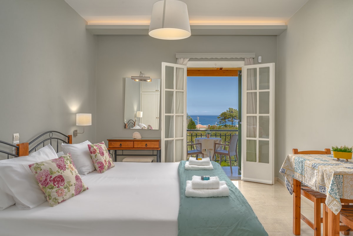 A spacious room features a large bed adorned with floral pillows and a mint green throw. Natural light floods in through open French doors, revealing a view of the sea and surrounding greenery. A wooden dresser and dining table enhance the welcoming ambiance.