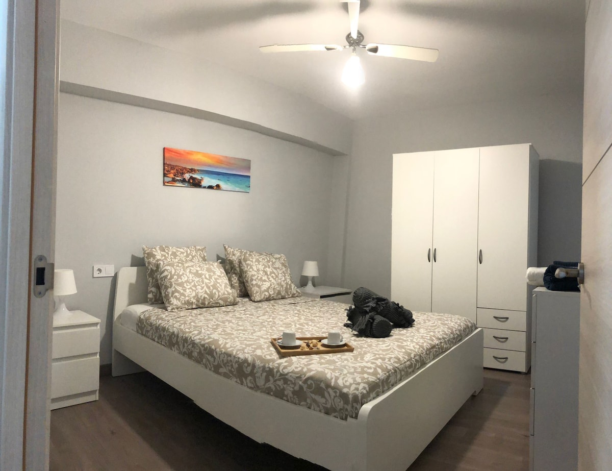 A serene bedroom is presented with a spacious bed adorned with patterned bed linens and soft pillows. Two nightstands, each with a lamp, flank the bed. A white wardrobe and a gray dresser are visible, while a ceiling fan contributes to the air circulation.
