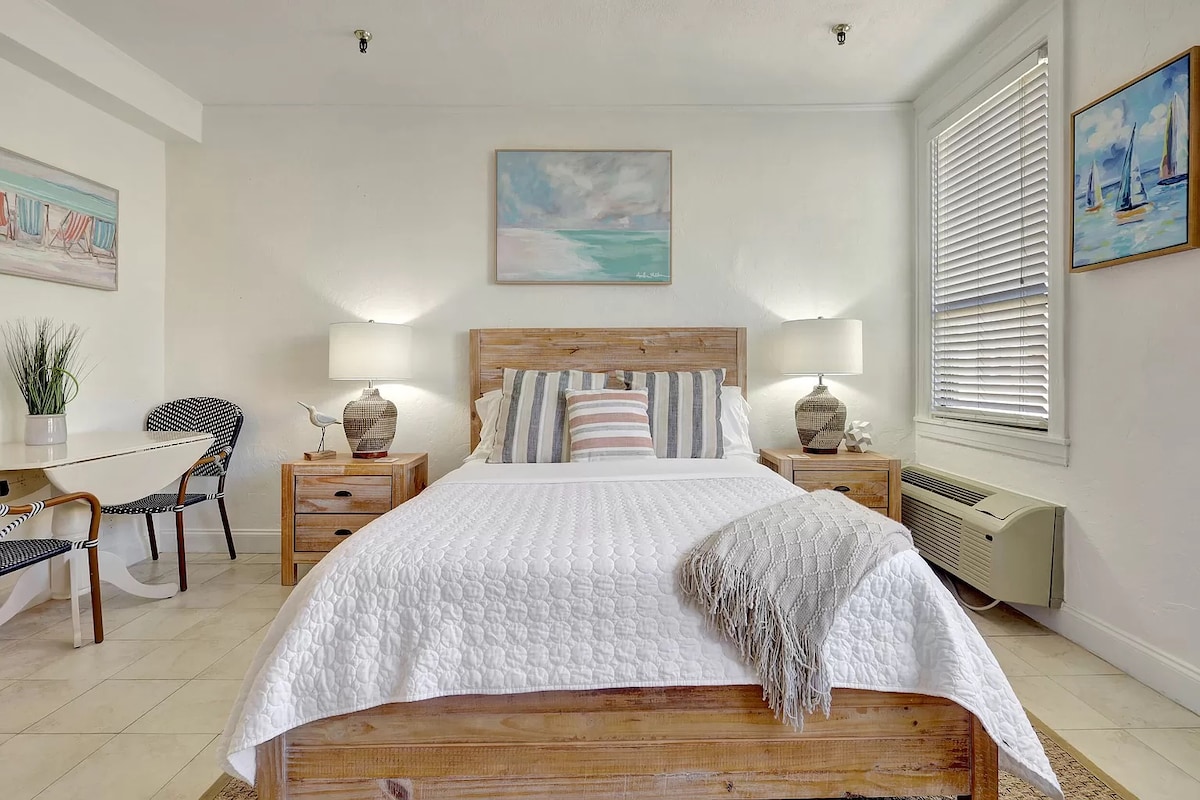 A cozy sleeping area features a queen bed with a textured white blanket and decorative pillows. Nightstands with lamps are positioned on either side. An adjoining dining space with a small table and chairs is visible, along with artwork depicting beach scenes and boats.