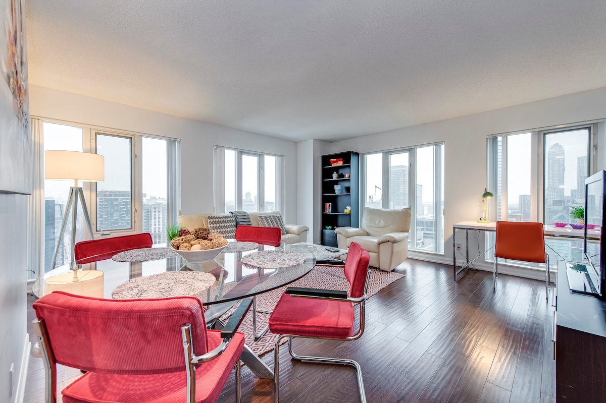 An open living and dining area is bathed in natural light from multiple windows. A round dining table with plush red chairs is positioned near a cozy seating area featuring cream-colored sofas, while a flat-screen TV is visible in the corner.