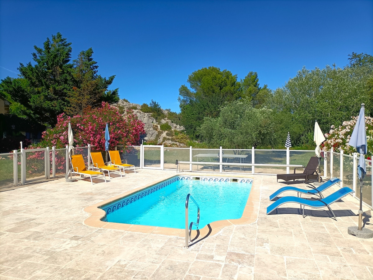 A sunlit pool area features a refreshing blue swimming pool surrounded by a spacious sun deck. Lounge chairs in various colors are positioned around the pool, and vibrant greenery and flowering plants create a scenic backdrop.