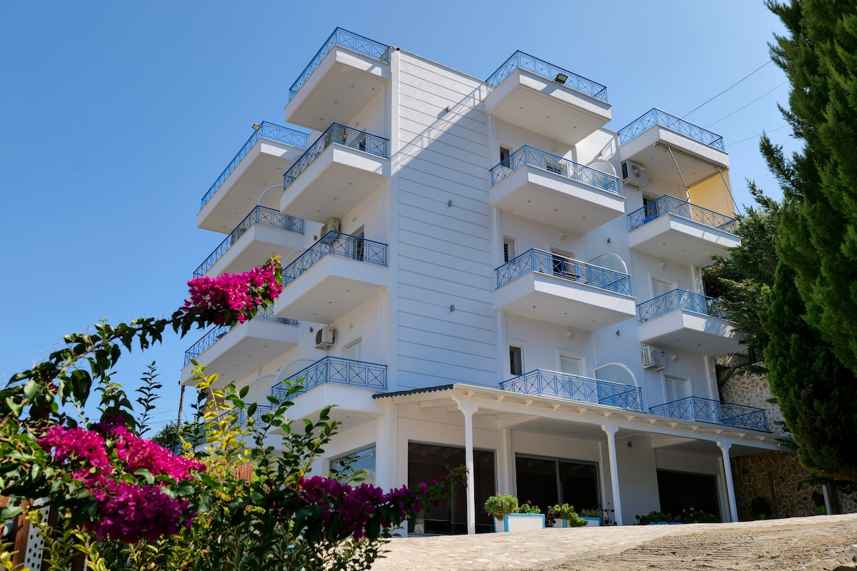 A multi-story building is presented, featuring multiple balconies with blue railings. The structure is framed by lush greenery and colorful flowering plants. The sunny sky enhances the building's fresh and modern design, indicating a welcoming atmosphere near the entrance.