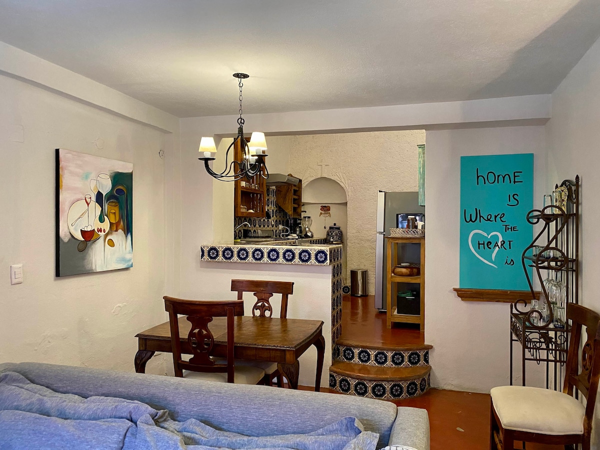 A comfortable living area features a dining table set for four and a spacious sofa. Colorful artwork adorns the walls, while a unique chandelier illuminates the space. A kitchen area is visible through an archway, complemented by traditional tile work.