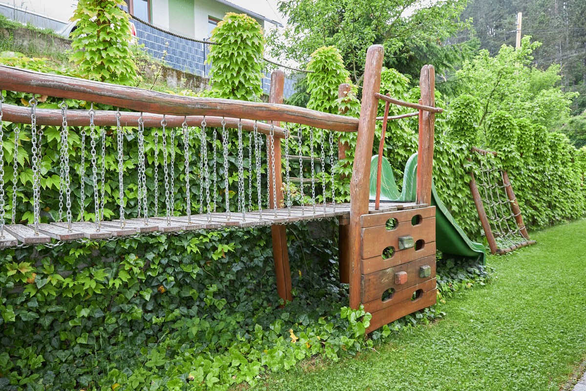 A wooden playground structure is seen nestled among vibrant green ivy. A swing set and climbing frame are positioned nearby, providing play options for children. The grassy area is lush and well-maintained, creating a natural and inviting environment.