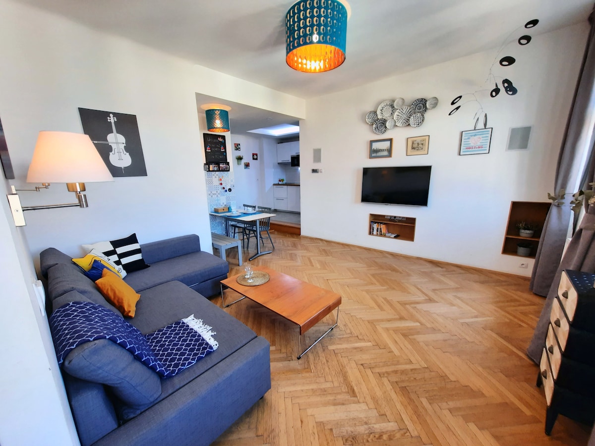 A light-filled living area features a comfortable blue sectional sofa with decorative pillows. A wooden coffee table sits in front of the sofa. A flat-screen TV is mounted on the wall opposite the seating, and a dining space is visible in the background.
