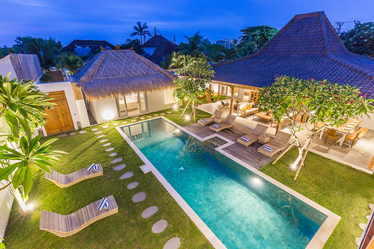 The image captures a serene villa at dusk, featuring a private swimming pool surrounded by lush greenery. Wooden lounge chairs are positioned along the poolside, with stepping stones leading through the garden. Traditional thatched roofs add to the tranquil atmosphere.