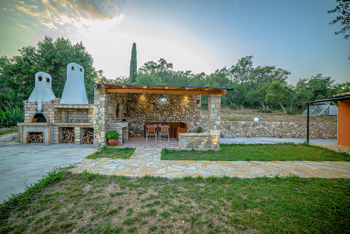 An outdoor stone kitchen area is showcased, featuring two traditional ovens and a wooden dining table with accompanying chairs. The space is surrounded by lush greenery, creating a serene setting for al fresco dining. A pathway is visible leading towards the area.