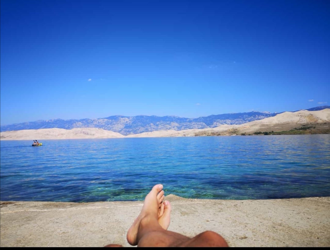 Clear blue waters are visible in the foreground, with a person’s feet resting on a rocky ledge. The background features gentle hills and mountains under a bright blue sky, creating a tranquil setting for relaxation.