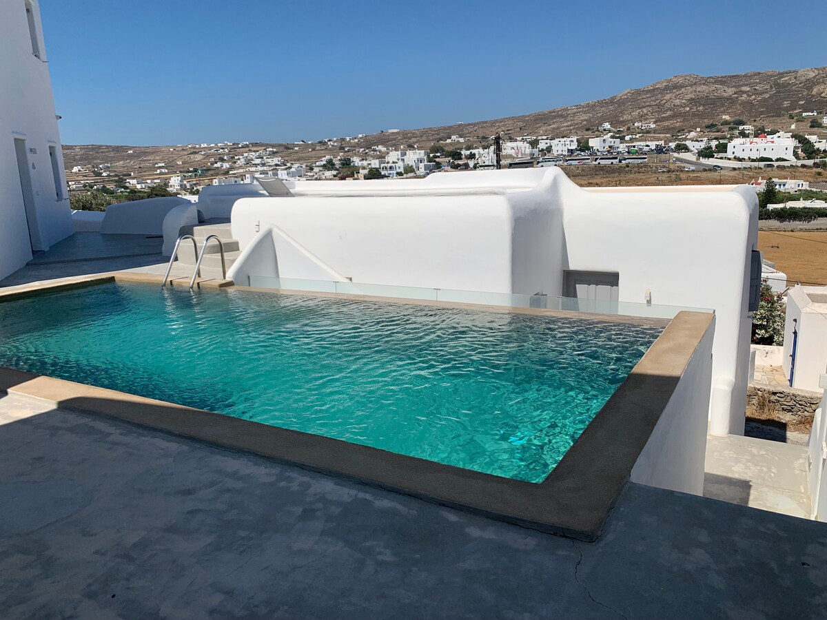 An inviting swimming pool is featured in this image, surrounded by a smooth concrete area. The pool's clear water reflects the sky, and the scenic view of traditional white buildings and distant hills can be seen in the background, providing a serene atmosphere.