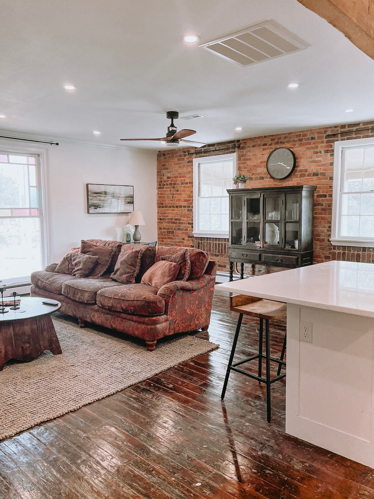 The living space features exposed brick walls and hardwood flooring, creating a warm ambiance. A large couch adorned with decorative pillows is positioned near a contemporary coffee table. Natural light enters through windows, while a bar area with stools adds to the inviting layout.