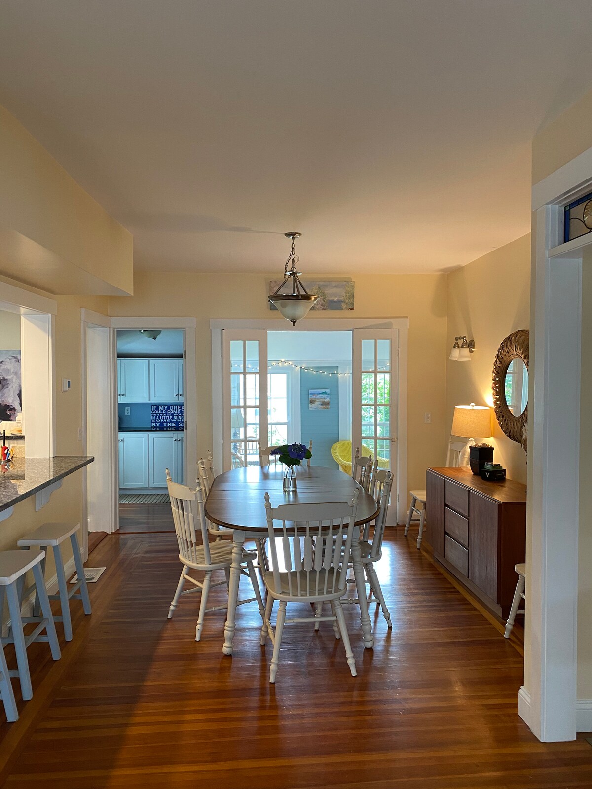 A dining area features a large round table surrounded by eight white wooden chairs. Natural light fills the space through multiple windows, illuminating the warm wood flooring. A console table holds a lamp, and a decorative mirror adds depth to the room.