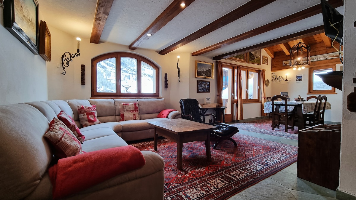 A cozy living area features a spacious sofa adorned with warm textiles, complemented by a large coffee table on a patterned rug. Natural light filters through multiple windows, enhancing the rustic decor on the walls. An adjacent dining area is visible, showcasing a table and chairs.
