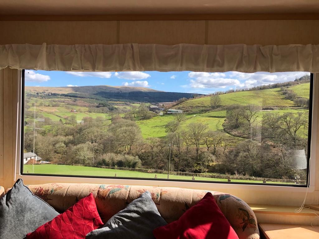A large window frames a view of rolling green hills and distant mountains under a bright blue sky. Soft plush cushions are arranged on a cozy seating area, inviting relaxation while enjoying the scenic landscape.