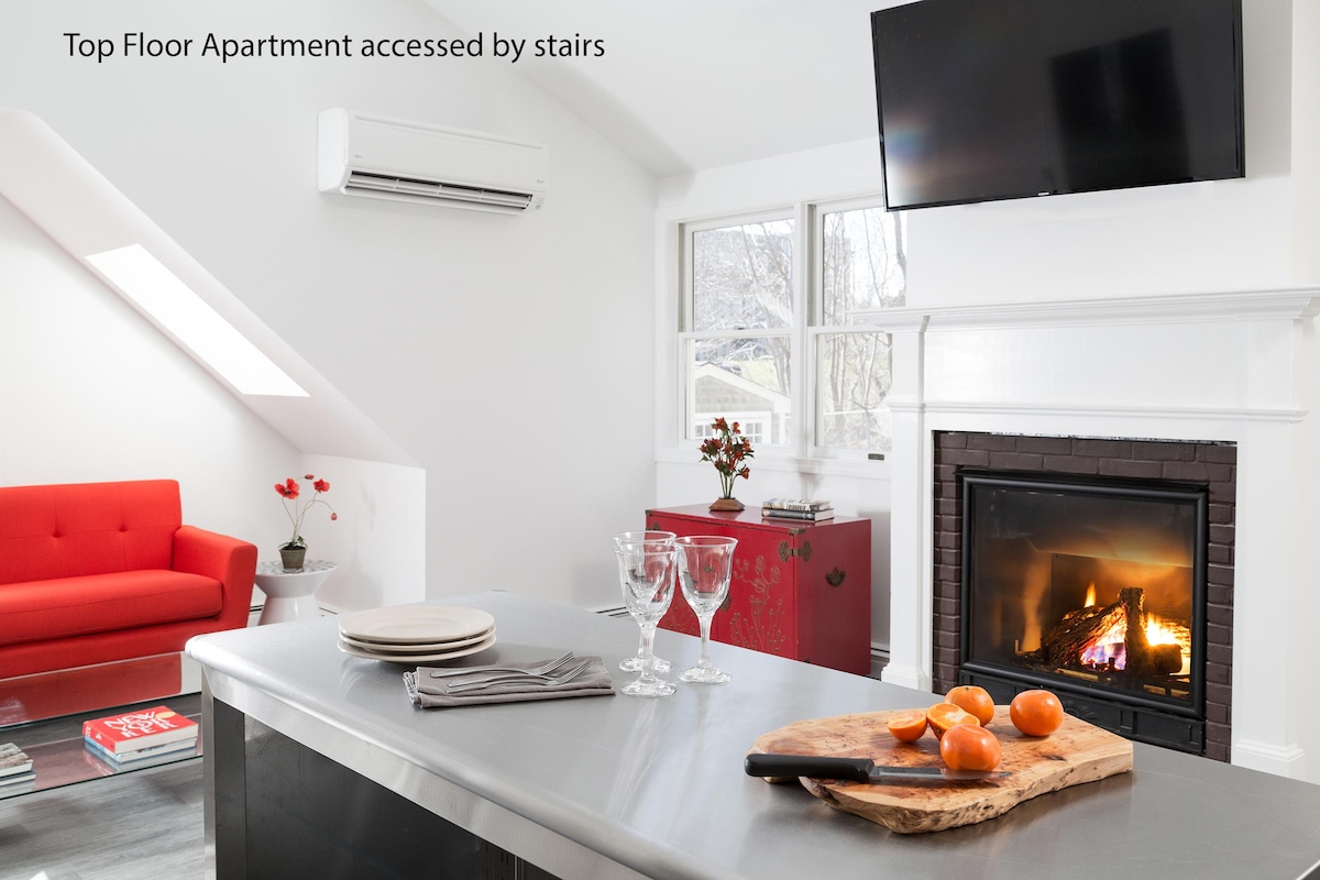 A bright living area is illuminated by natural light, featuring a modern red sofa beside a fireplace. An elegant dining space is highlighted by a polished countertop and wine glasses. A flat-screen TV is mounted above the fireplace, enhancing the contemporary decor.