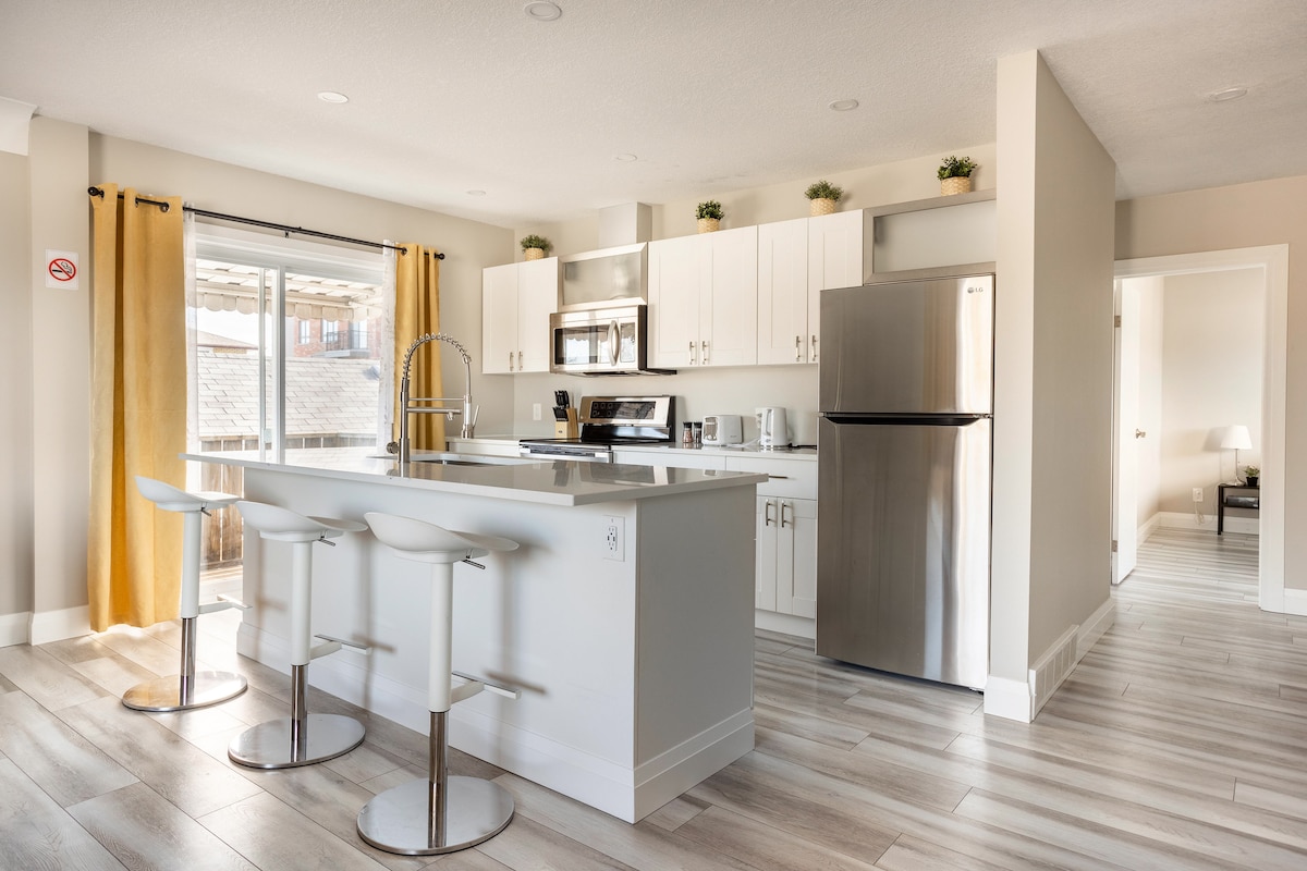 A modern kitchen features stainless steel appliances and white cabinetry. A central island with three bar stools provides additional seating. The bright space is illuminated by natural light through sliding glass doors, which lead to an outdoor area, complemented by yellow curtains framing the windows.
