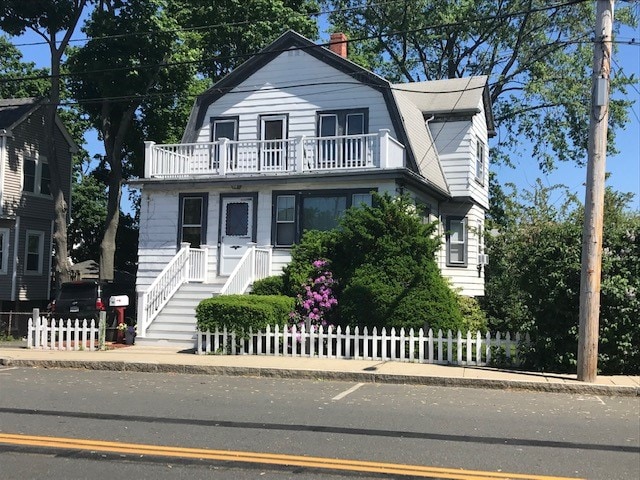 A two-story house with a light blue exterior features a white railing balcony on the second floor. Lush greenery and flowering plants surround the property. A white picket fence encloses the front yard, while clearly defined pathways lead to the entrance. The clear blue sky provides a bright backdrop.