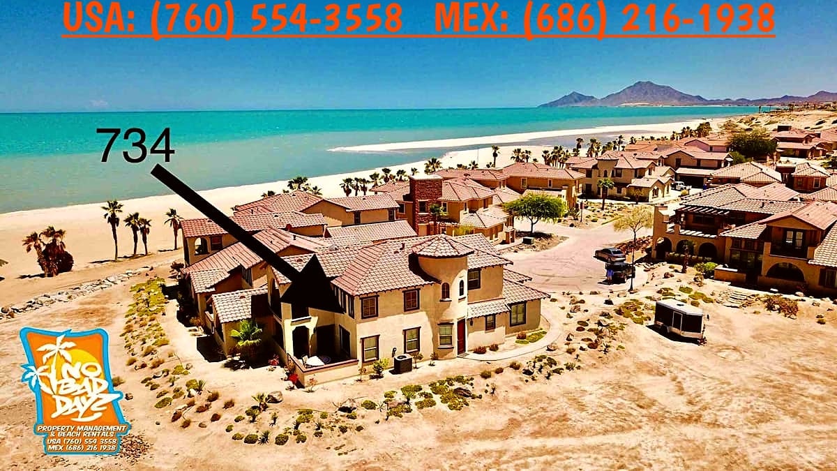 An aerial view showcases a two-story condo at La Ventana del Mar Beach and Golf resort, surrounded by landscaped areas and palm trees. The turquoise waters of the Sea of Cortez are visible in the background, enhancing the coastal setting. Nearby residential structures are also seen.
