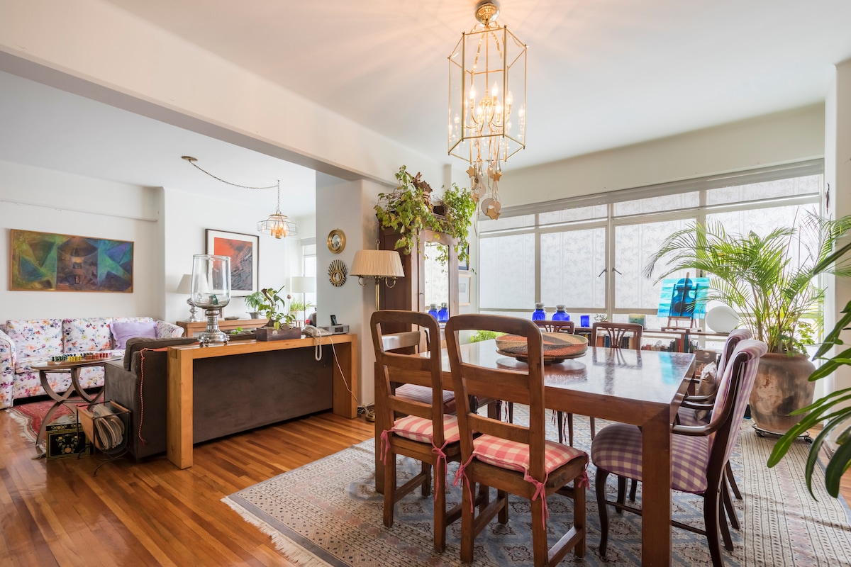 An open living area features a large wooden dining table surrounded by various wooden chairs. Natural light filters through expansive windows adorned with delicate curtains. A colorful, patterned sofa is visible in the background, along with decorative plants enhancing the space.