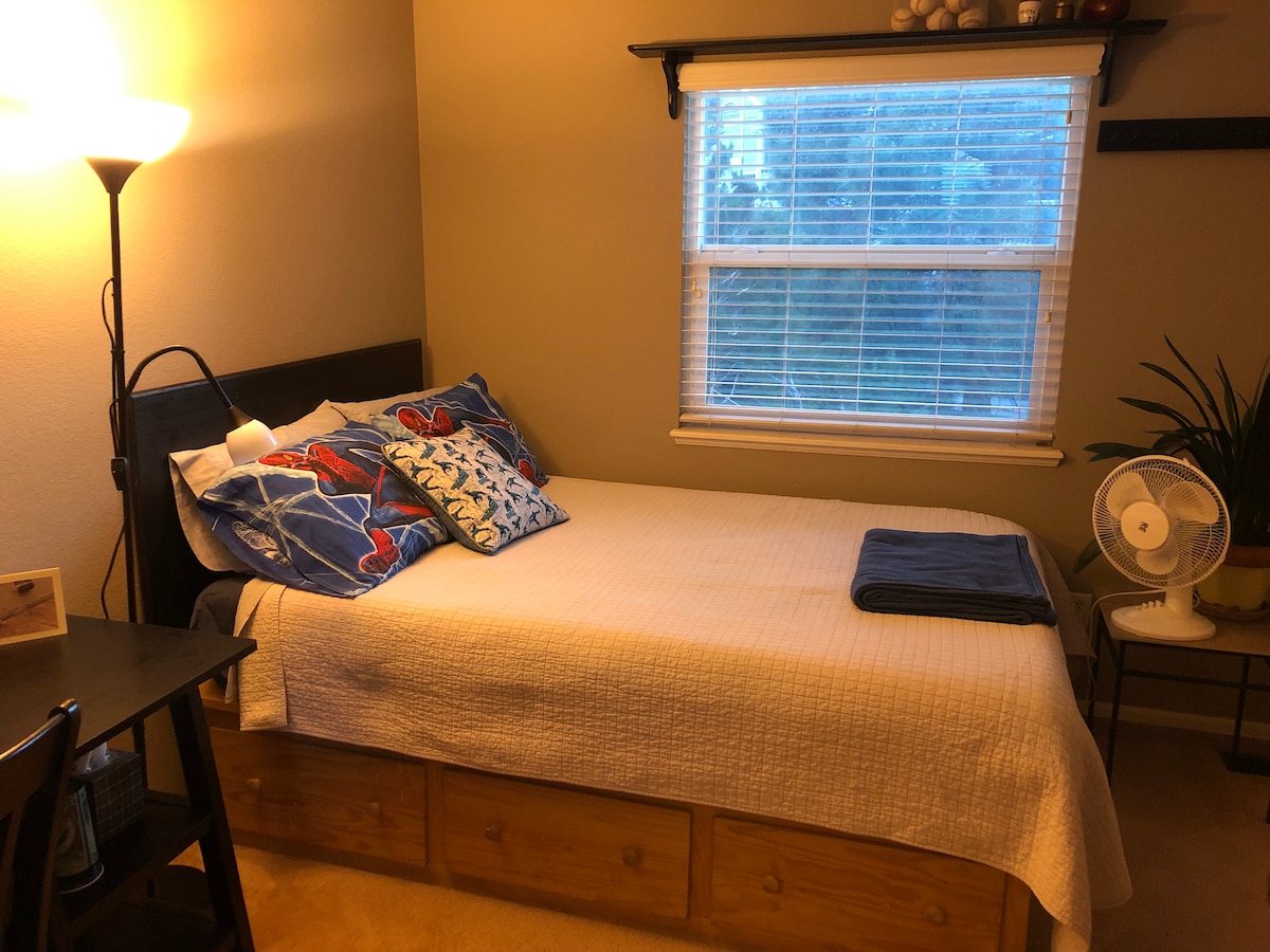 A small bedroom is presented with a full-size bed adorned with a quilt. A decorative lamp and a desk are located beside the bed. Light filters through a window covered by blinds. A plant and a fan are positioned nearby on a stand.