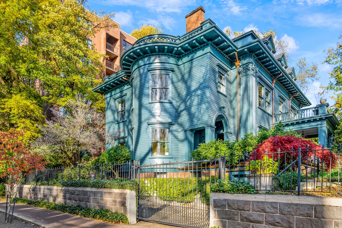 The historic Isam White House features a striking blue exterior with intricate details and large windows. Lush greenery surrounds the property, complemented by seasonal foliage. The well-maintained garden is enclosed by a decorative black iron fence, adding to the home's charm.