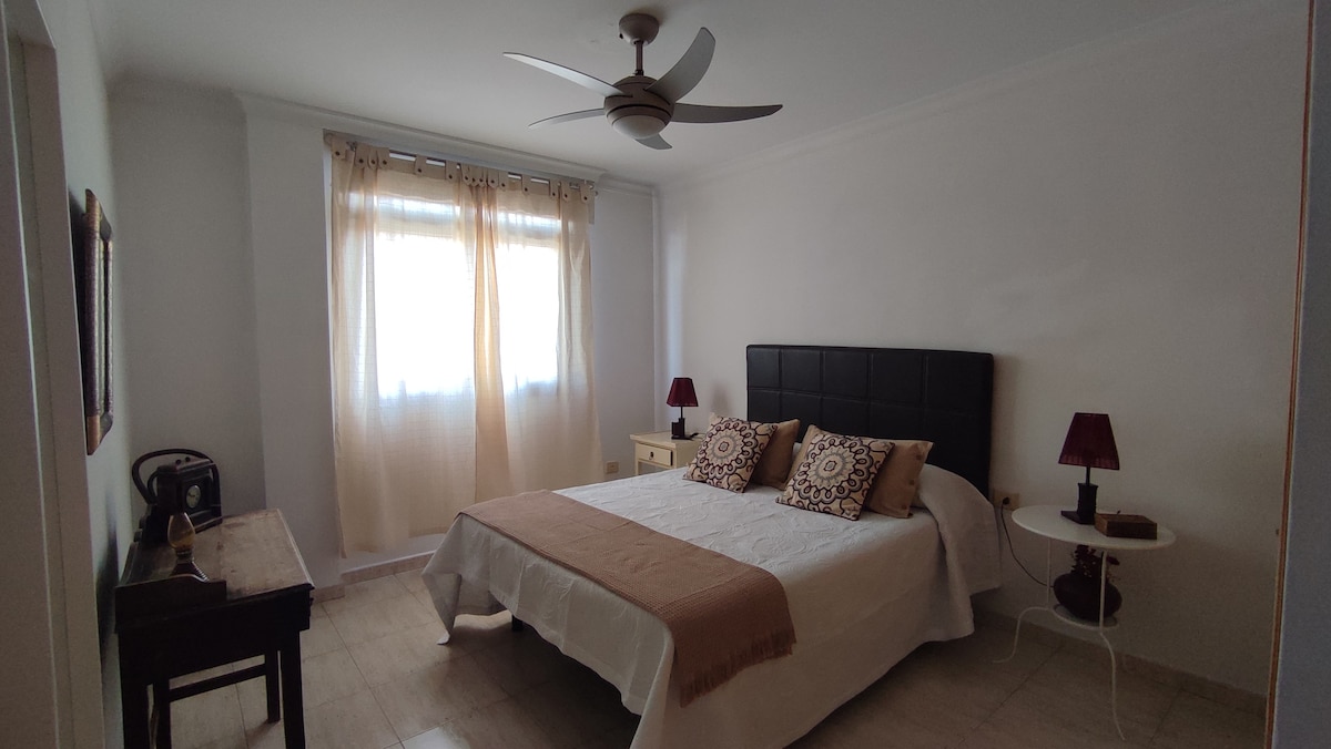 A bright bedroom features a spacious bed with dark upholstered headboard and decorative pillows. Two bedside tables with lamps are placed on either side. Natural light filters through sheer curtains, and a ceiling fan adds to the room's comfort. A wooden writing desk is positioned against the wall.