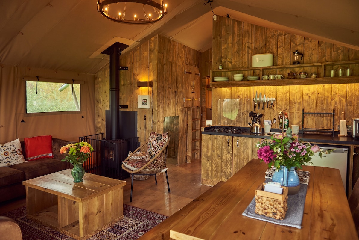 An inviting living area is shown with a wooden dining table at the forefront and a cozy sofa accompanied by a patterned chair. A log burner stands beside the rustic wood cabinetry, where kitchen essentials are neatly arranged. Fresh flowers adorn the table, adding a vibrant touch.