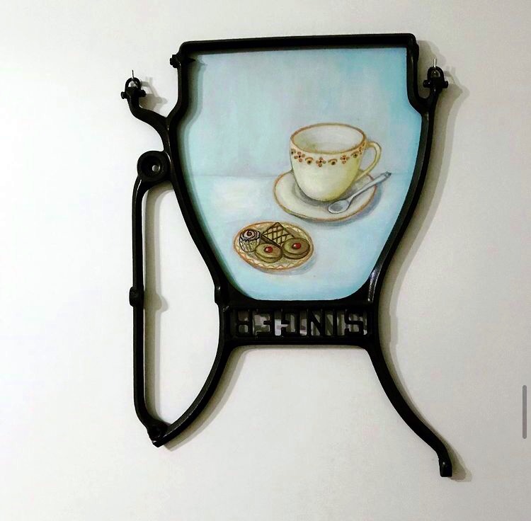 A whimsical wall art piece featuring a painted cup and a plate of cookies. The design is framed by a decorative, black metal structure that resembles a vintage chair. Soft colors in the artwork create a light and airy feel.