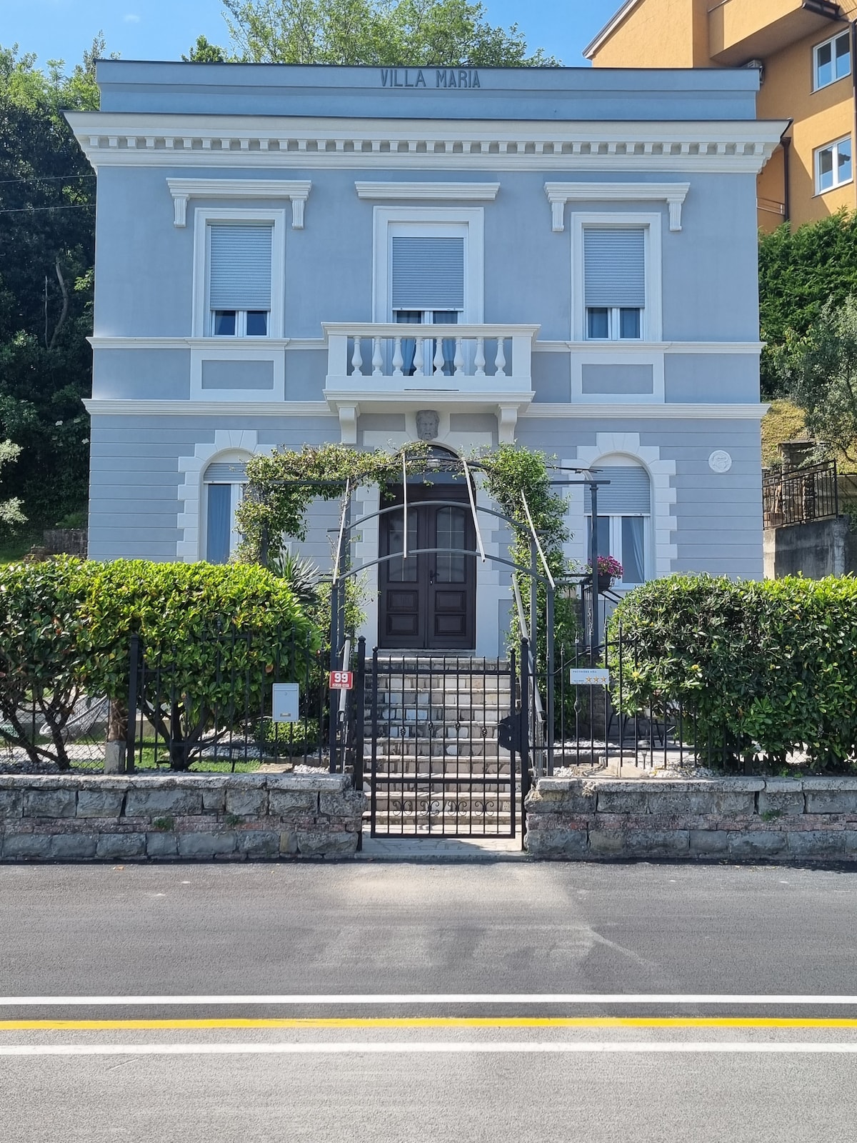 The elegant gray building features a classic façade with intricate architectural details. A gated entrance is surrounded by well-maintained hedges and greenery. A series of steps lead up to the front door, framed by a small balcony and ornate features above.