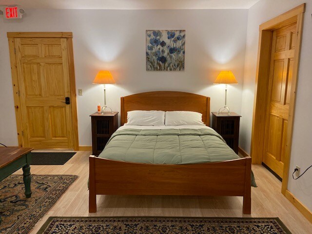 The guest suite features a spacious queen bed framed with natural wood and dressed in light linens. Two bedside lamps provide soft illumination, while artwork adorns the wall above the bed. A wooden door leads to the hallway, and a sturdy dresser stands beside the bed.