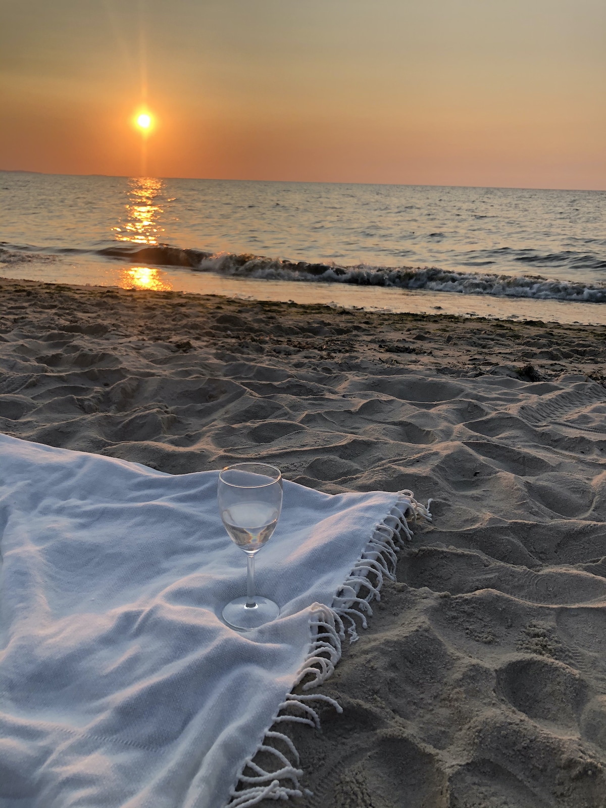 A serene beach scene is captured at sunset, with gentle waves lapping at the shore. A white blanket is spread on the sand, and a glass is placed on it, reflecting the soft golden hues of the setting sun.