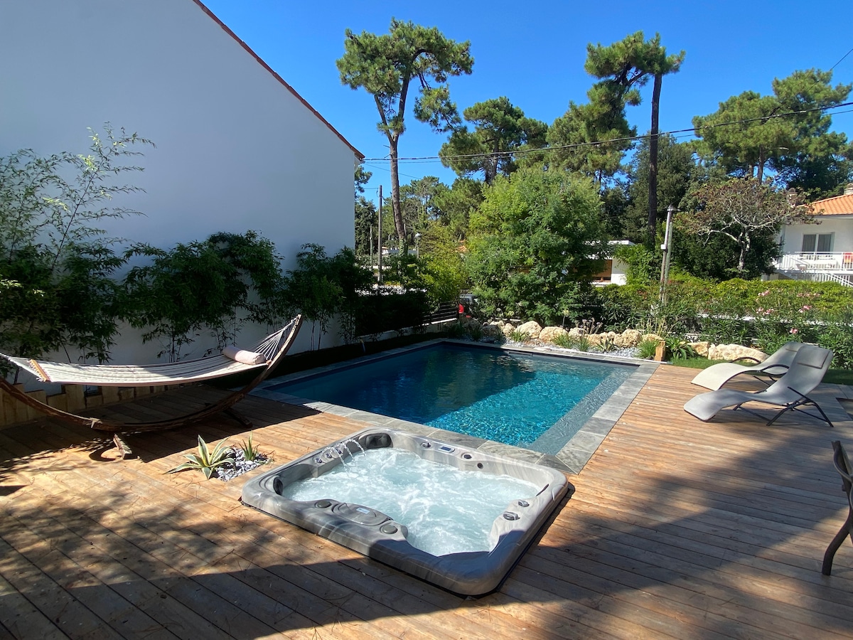 A private deck features a relaxing jacuzzi and a swimming pool surrounded by wooden decking. A hammock is situated nearby, framed by lush greenery and tall pine trees. The clear blue sky above enhances the serene outdoor space.