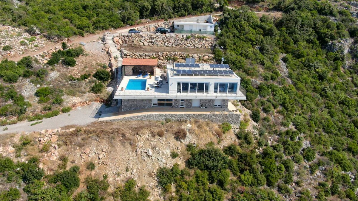 An aerial view captures a modern eco-friendly house positioned on a hillside, surrounded by lush greenery. The home features a solar panel roof and a private pool on the terrace. The landscaped area above rocky terrain creates a natural, serene setting.
