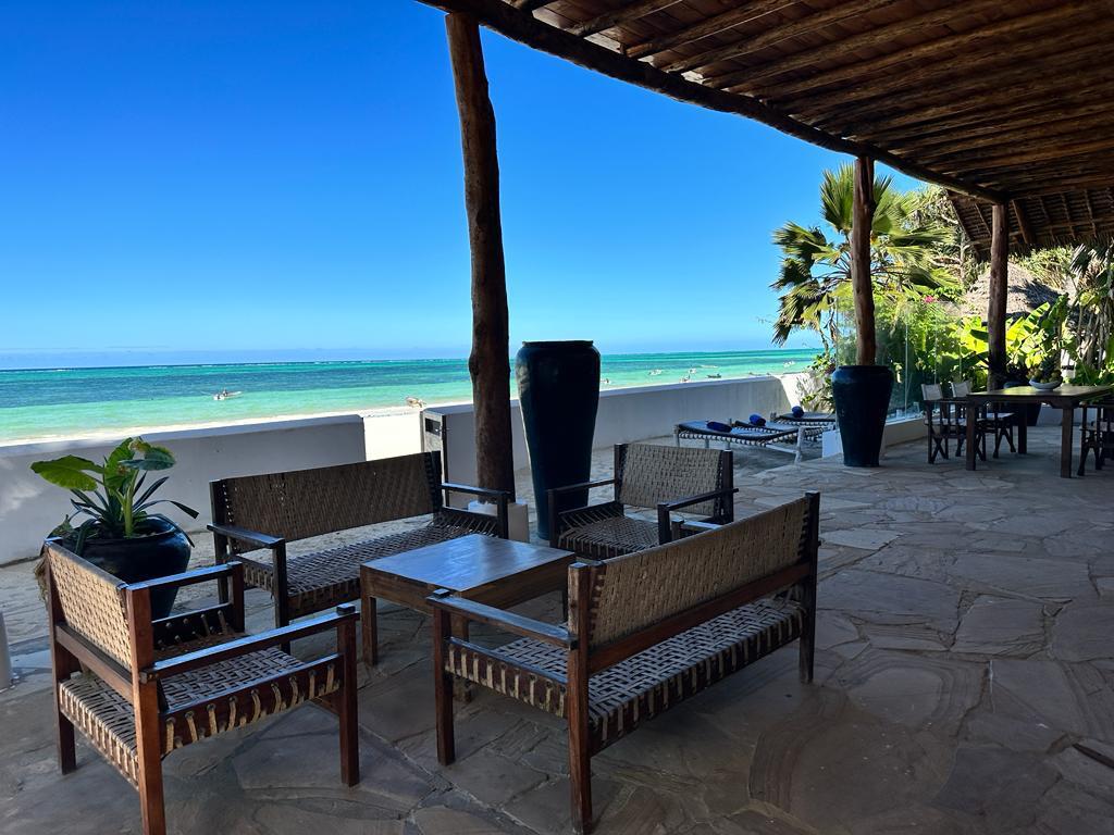 A covered patio area features natural wood beams and stone flooring. Comfortable seating is arranged around a wooden table, complemented by potted plants. The ocean view showcases turquoise waters and a clear blue sky, enhancing the ambiance of the space.