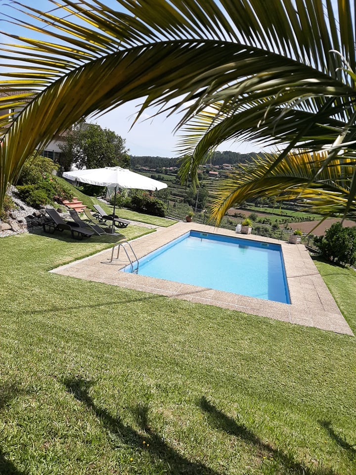 A private swimming pool is framed by a well-maintained lawn, with sun loungers positioned alongside. A large umbrella provides shade, while a scenic view of rolling hills and greenery is visible in the background.