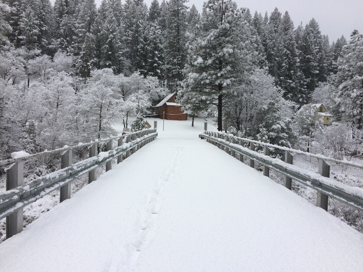 A snow-covered bridge extends through a serene landscape, surrounded by densely packed evergreen trees. A quaint cabin is visible in the background, partially obscured by the frost-covered branches. Footsteps in the fresh snow lead across the bridge, inviting exploration of the winter wonderland.