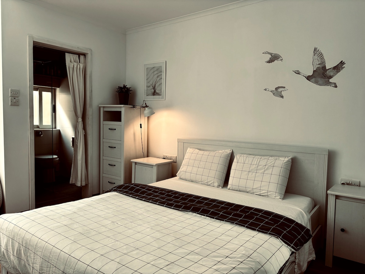 A bright bedroom features a king-sized bed with a grid-patterned duvet and two accent pillows. A bedside lamp illuminates the space, while a wall decoration of flying geese adds an artistic touch. A door leads to a small bathroom, partly visible in the background.